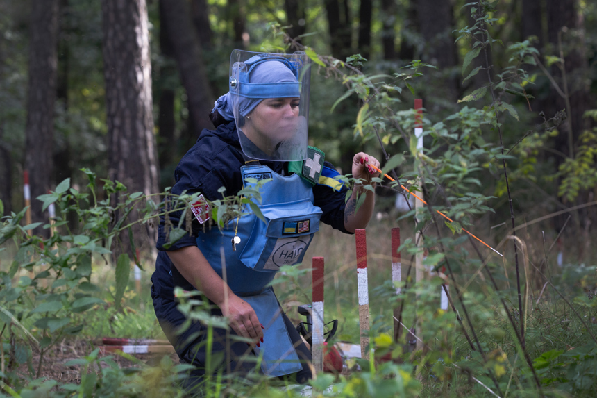 Karina Krasnikova, a female deminer from Trostianets Village, kneels in front of mine marking sticks 