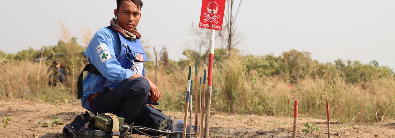 A deminer crouches next to a mine warning sign with his PPE and metal detector next to him