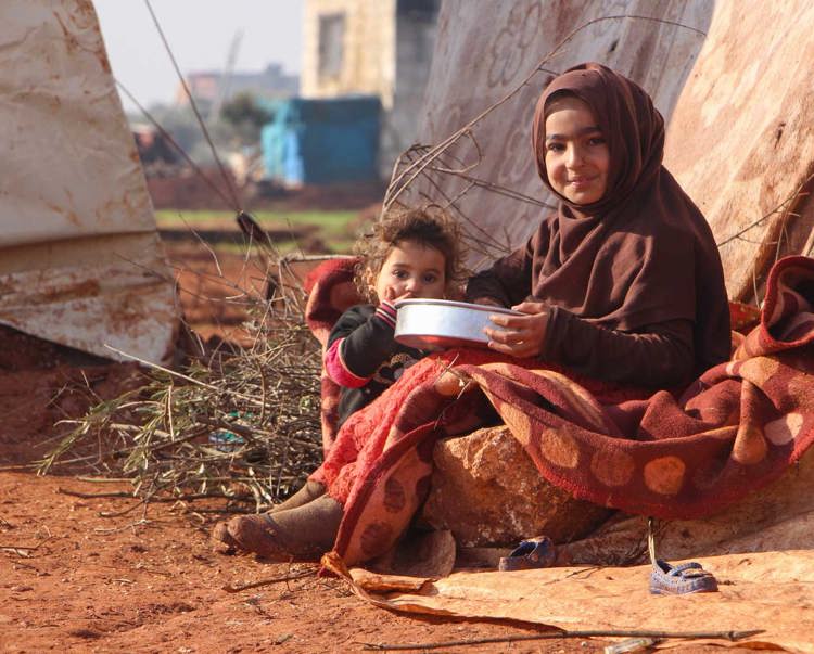 A child and a toddler sit on the ground holding a metal bowl