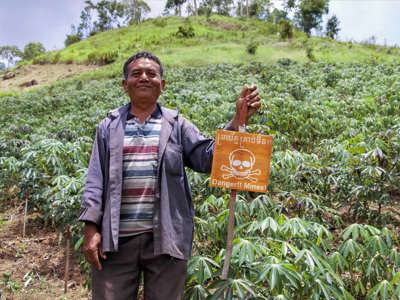 Pork Chei stands next to a mine warning sign in a Cambodian crop field