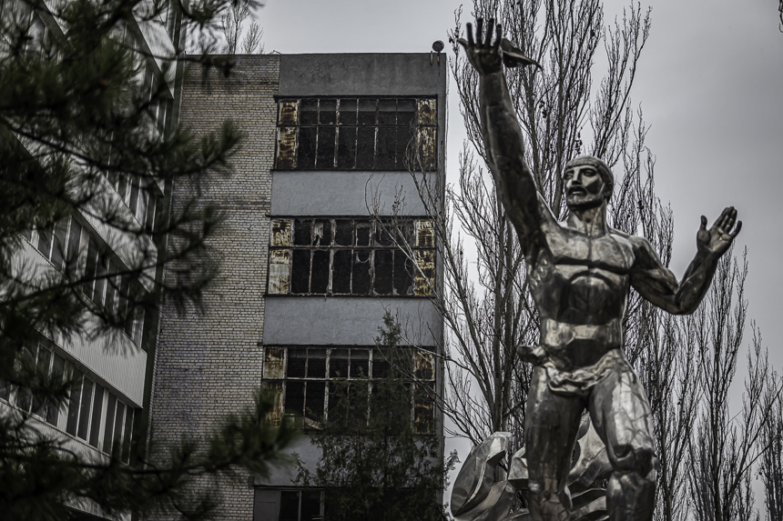 A silver statue of a man stands in the foreground of damaged buildings