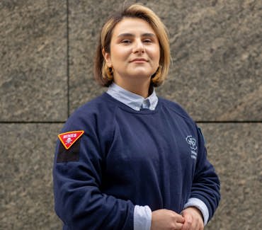 Портрет експертки з інформування про мінну небезпеку в синьому светрі зі знаком "MINES" на фоні стіни будівлі