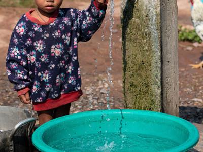 A little boy holds on to a tap running water into a bucket outside
