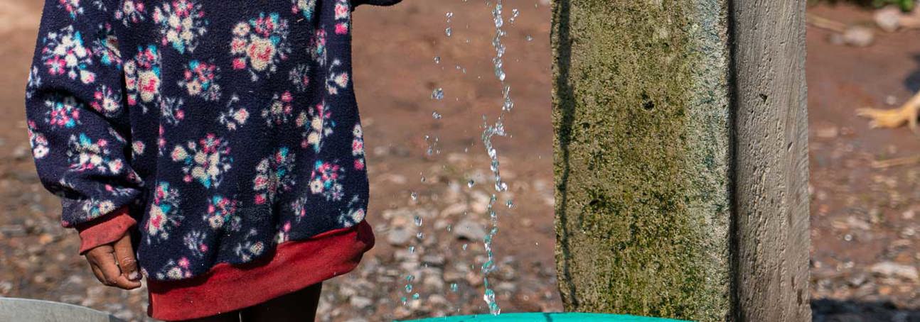 A little boy holds on to a tap running water into a bucket outside