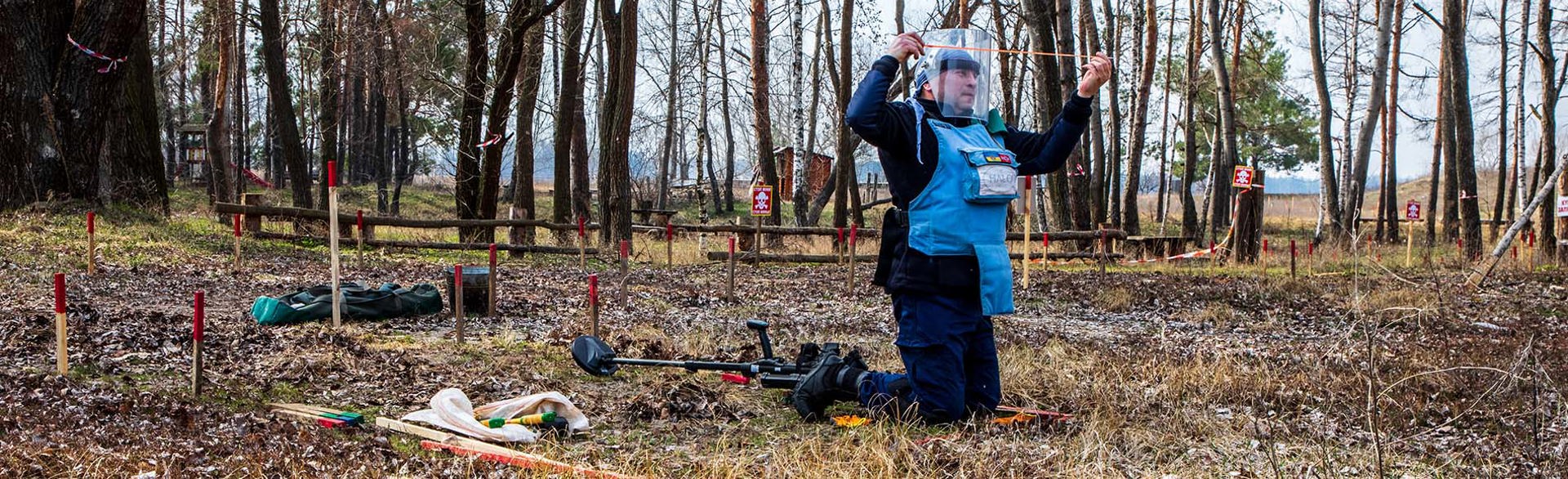 Демінер в захисному спорядженні з щупом в руках проводить пошук розтяжок на фоні інструментів та детектора в осінньому лісі