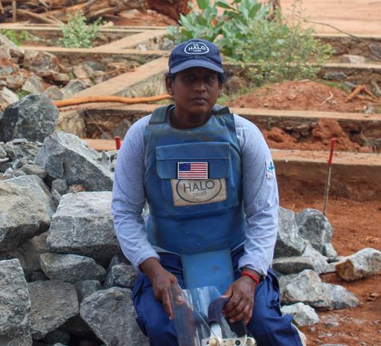 Sivaginedavi, a HALO deminer, sits on some rocks outside next to a mine marking stick