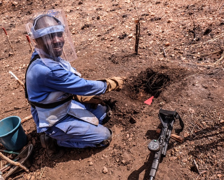 Lovemore Tarugarira, a deminer, kneels in the soil pointing to a hole he dug