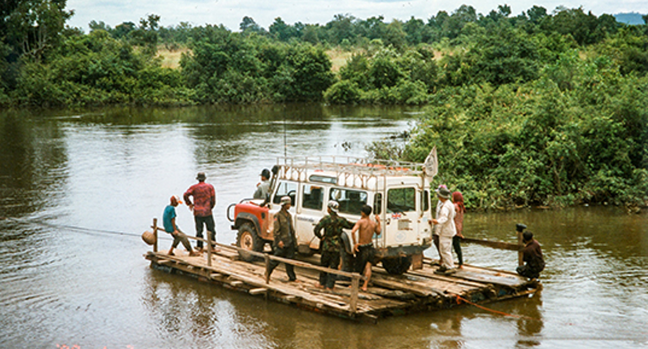 A large vehicle and group of people cross a river on rustic wooden boat in Cambodia
