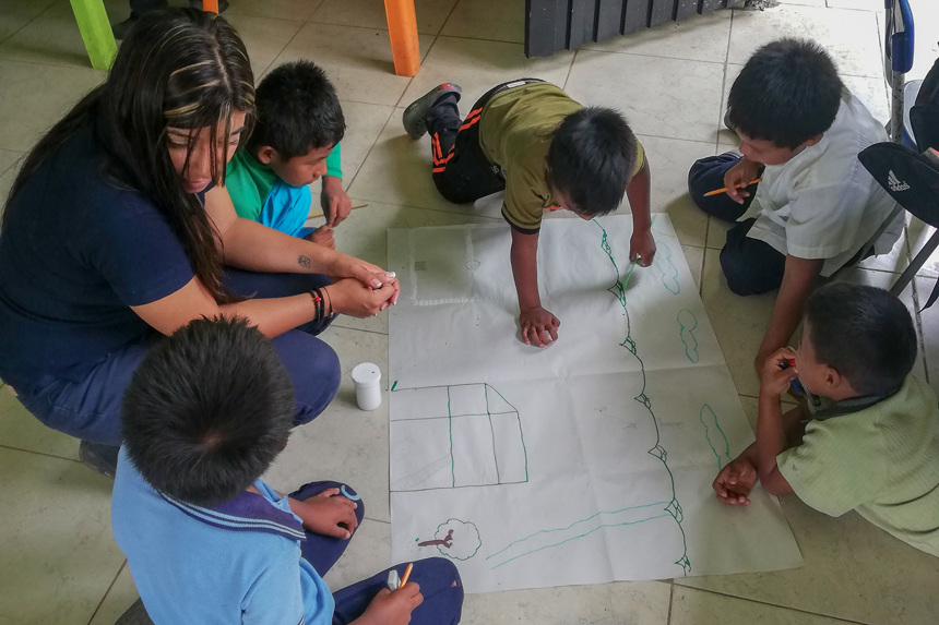 A woman and five children sit on the floor to draw on a large piece of paper