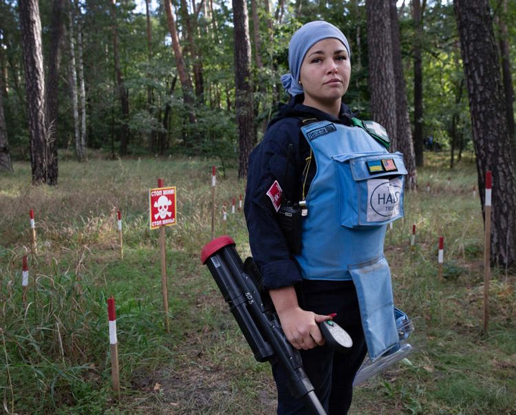 A woman stands tall in her demining gear while holding her metal detector in a minefield