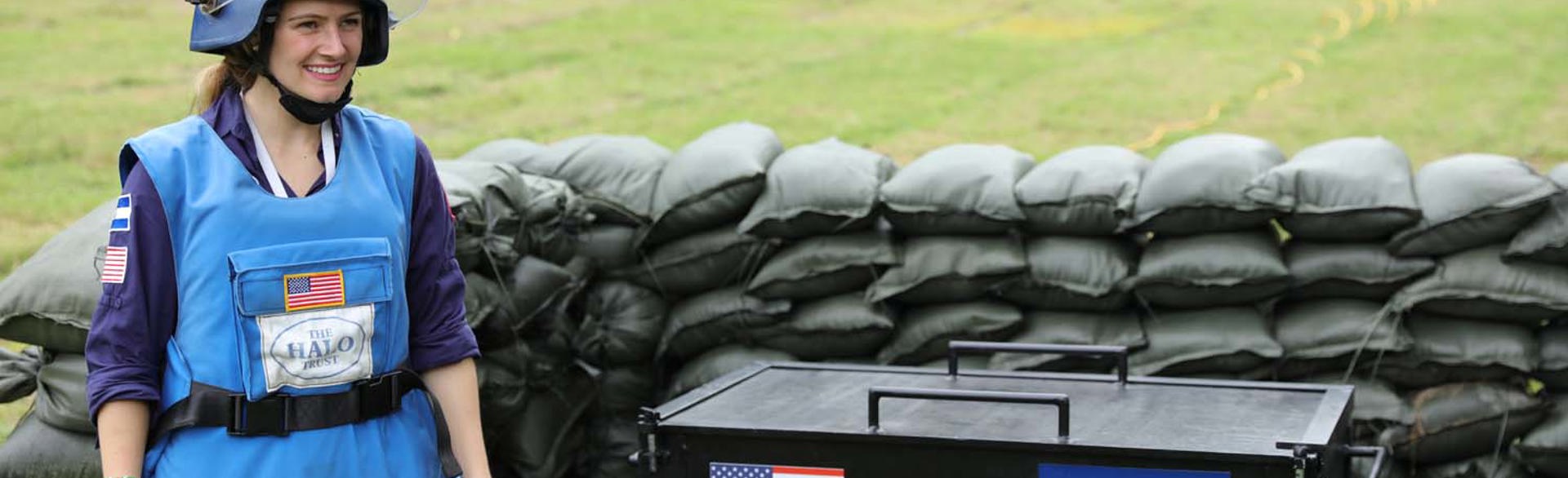 A deminer stands next to a large container with the HALO and US flag surrounded by sandbags 
