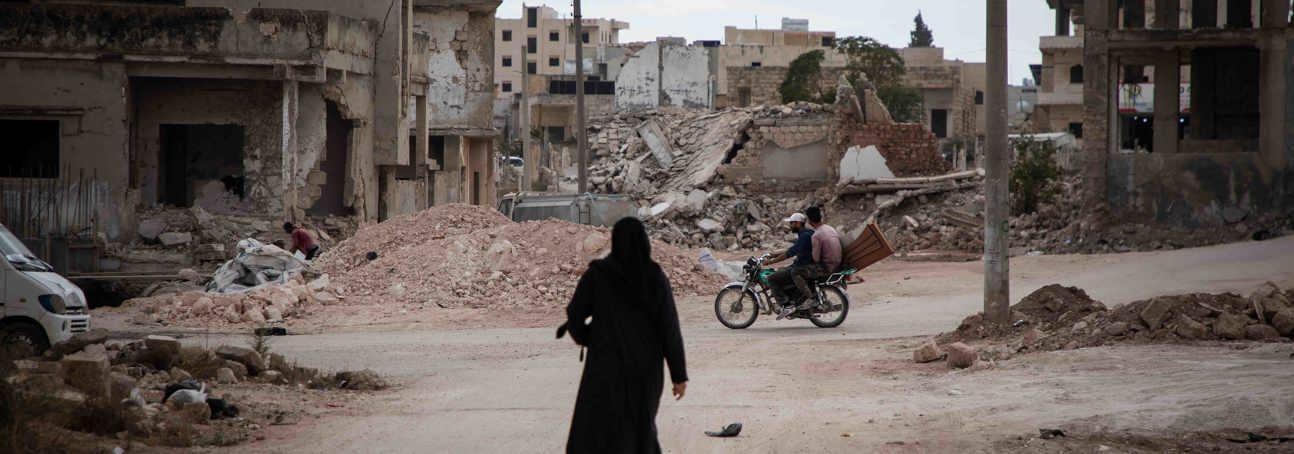 Woman dressed in black, walking through a street in Syria surrounded by rubble from destroyed buildings