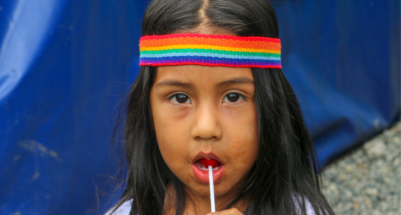 A child wears a rainbow bandana whilst eating a lolly pop