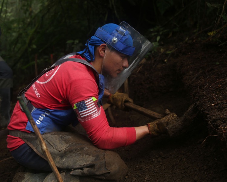 A deminer wearing PPE uses tools to dig at the soil