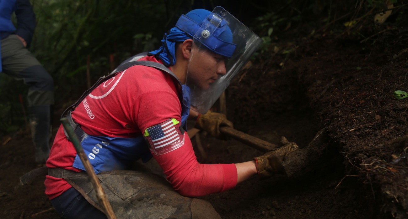 A deminer wearing PPE uses tools to dig at the soil