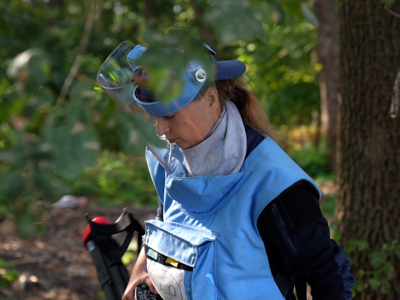 A deminer wearing PPE holds a metal detector in a forest