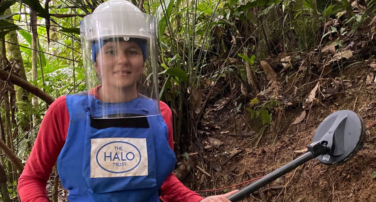 A female deminer wearing PPE holds a metal detector