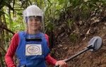 A female deminer wearing PPE holds a metal detector