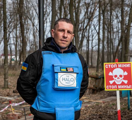 A deminer stands next to a mine warning sign with trees in the background