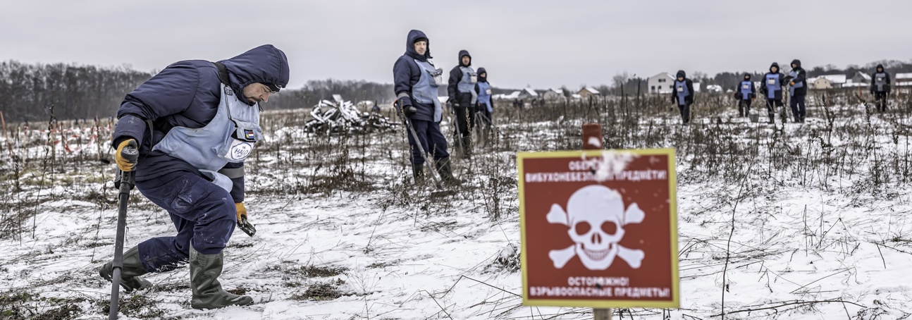 deminers wearing PPE use a metal detector in a snow covered minefield with mine warning  signs