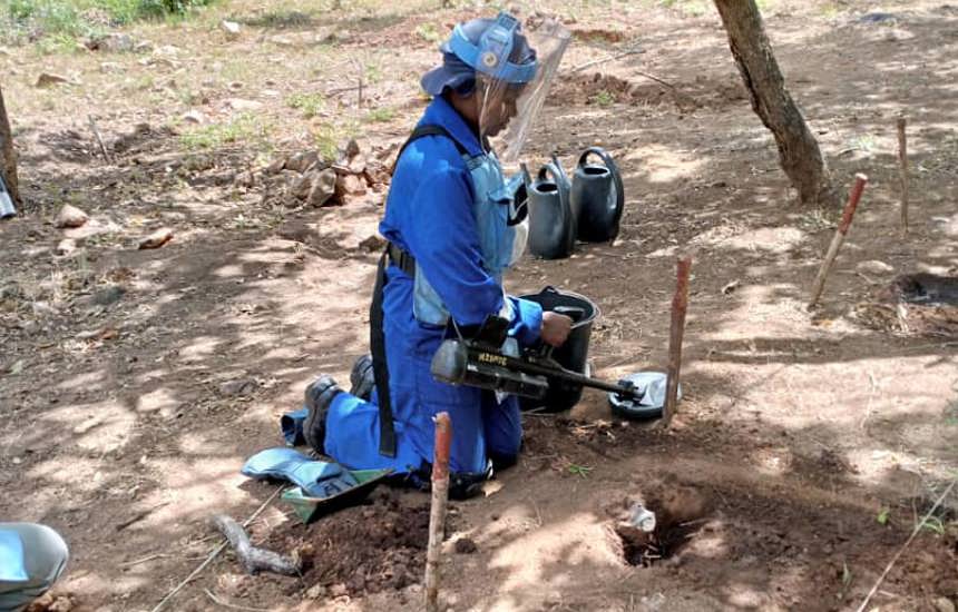 GIS specialist, Delight, wears PPE and holds a metal detector whilst kneeling to find mines in the ground 