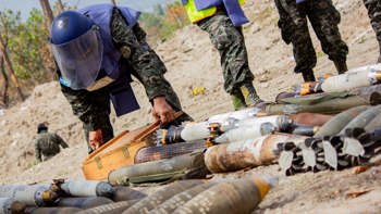 A person in a HALO vest and visor carefully handling a box among a stockpile of weapons in Honduras