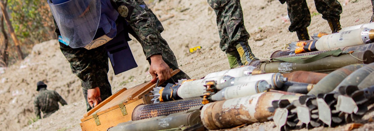 A person in a HALO vest and visor carefully handling a box among a stockpile of weapons in Honduras