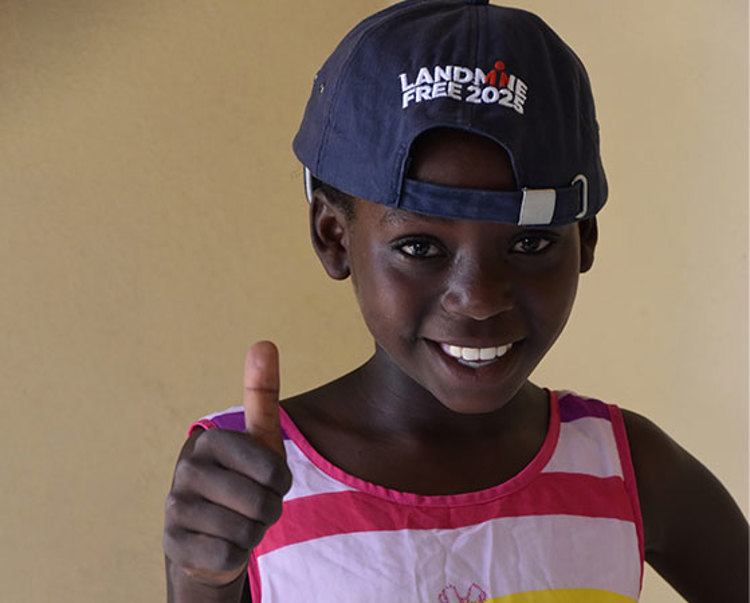 A boy poses with a thumbs up wearing a cap that says land mine free 2025