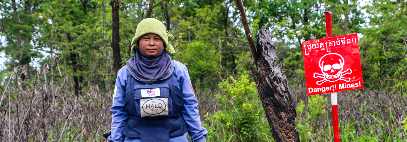 A deminer in Cambodia holds a detector and visor while walking through a minefield 