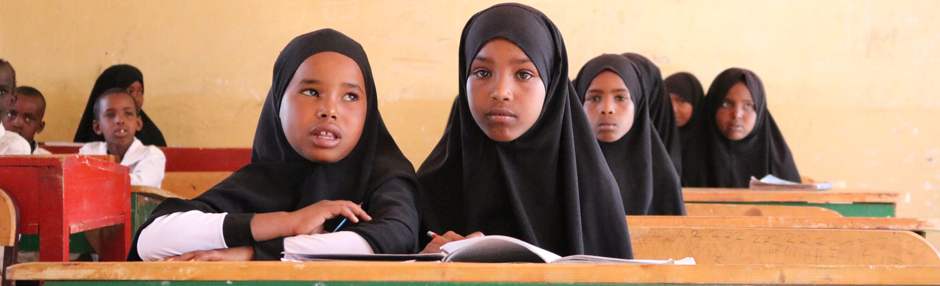 Two young girls sit in a classroom behind a desk that says "educate girls end poverty"