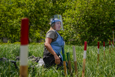 A deminer wearing PPE kneels in a field with mine marking sticks