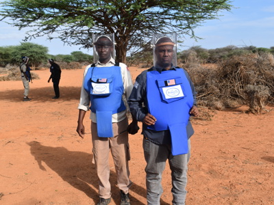 Two deminers pose together wearing their protective gear in a rural area