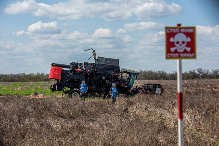 Deminers walk through a minefield with a mine warning sign and damaged farming vehicle