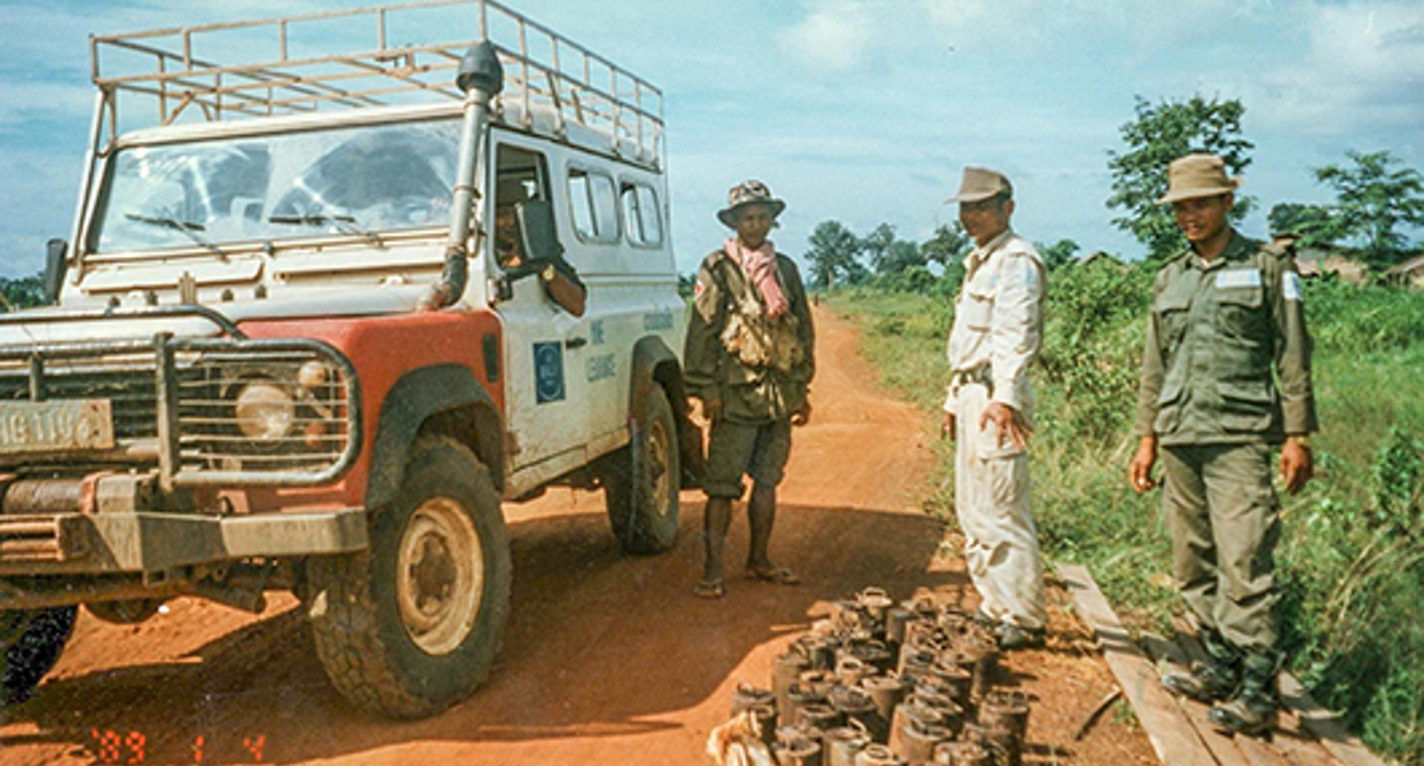 Three men stand next to a vehicle on a sandy track next to a large pile of mines