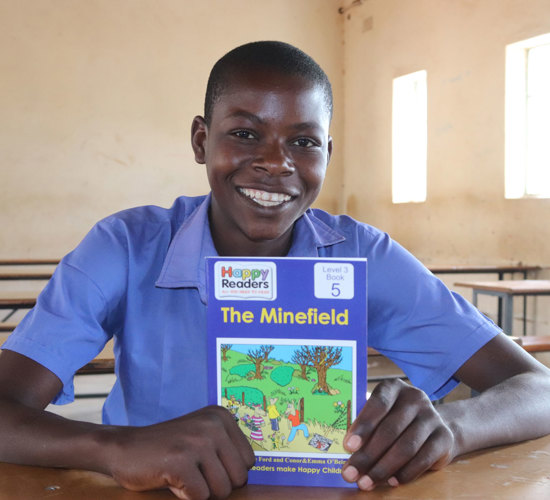A young boy in a class room smiles behind his desk with a landmine book