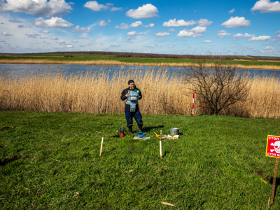 A HALO deminer stands in PPE with his equipment in a grassy minefield in front of a lake