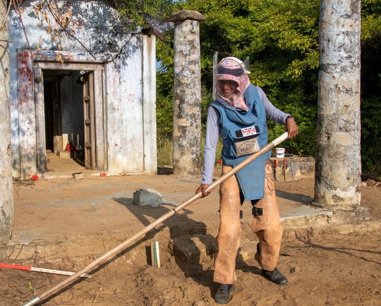 A deminer in PPE uses a rake on the soil in front of a destroyed building