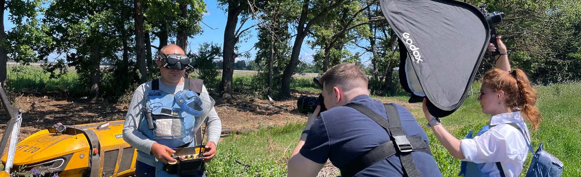 Журналісти фотографують демінера в захисному спорядженні з пультом керування від машини для дистанційної зрізки рослинності Robocut на фоні цієї машини біля лісопосадки