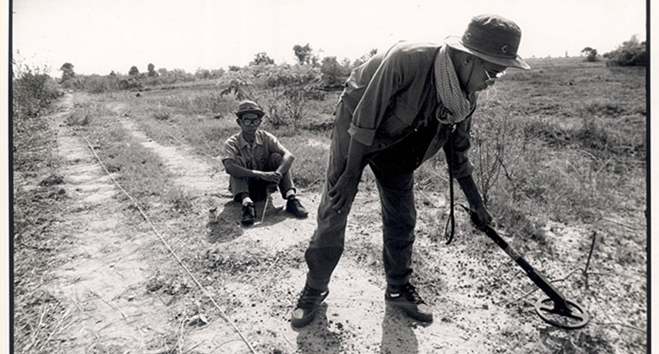 A black and white image of a deminer sitting next to another deminer using a detector on a track in Cambodia