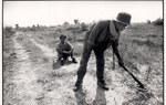 A black and white image of a deminer sitting next to another deminer using a detector on a track in Cambodia