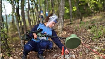 A deminer in PPE holds a metal detector and carries out demining activities on a hillside