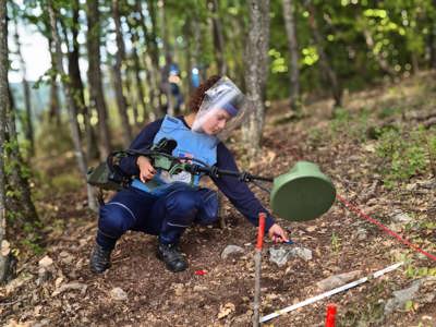 A deminer in PPE holds a metal detector and carries out demining activities on a hillside