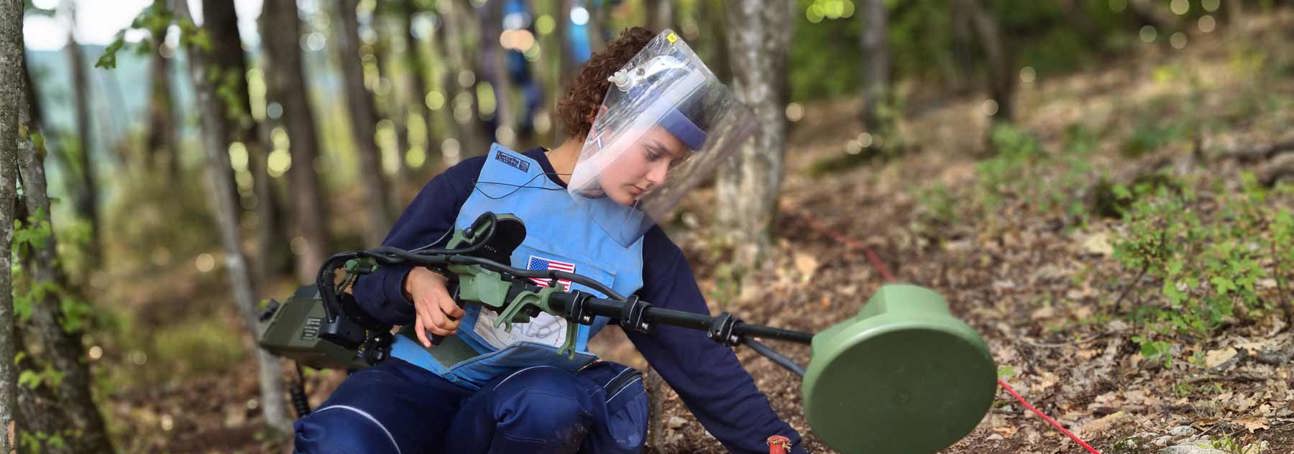 A deminer in PPE holds a metal detector and carries out demining activities on a hillside