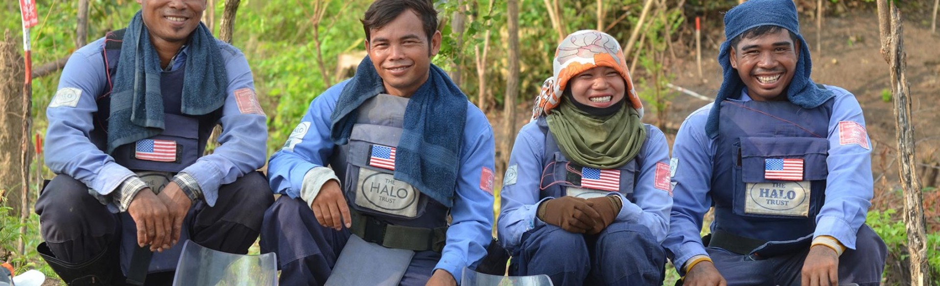 The demining team in Cambodia pose kneeling down and smile whilst wearing their PPE 