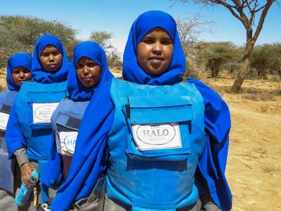 Four female deminers stand lined up in a rural area wearing PPE