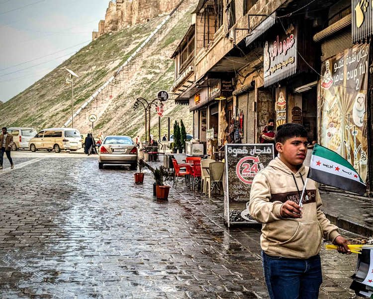 In Aleppo Citadel a boy stands by the side walk with a Syrian flag
