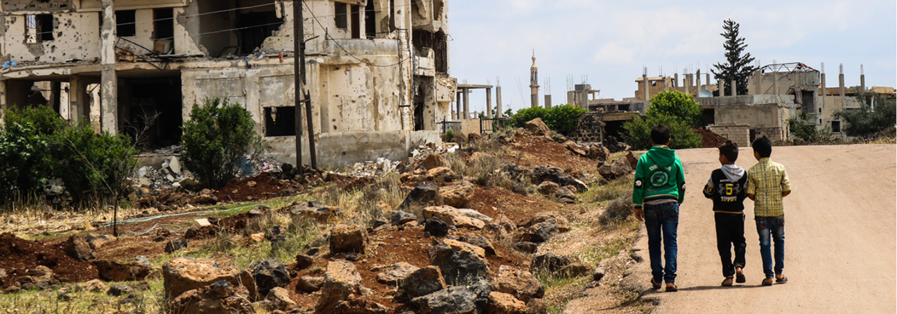 Three children walk on a road next to rubble and debris and destroyed buildings in Syria