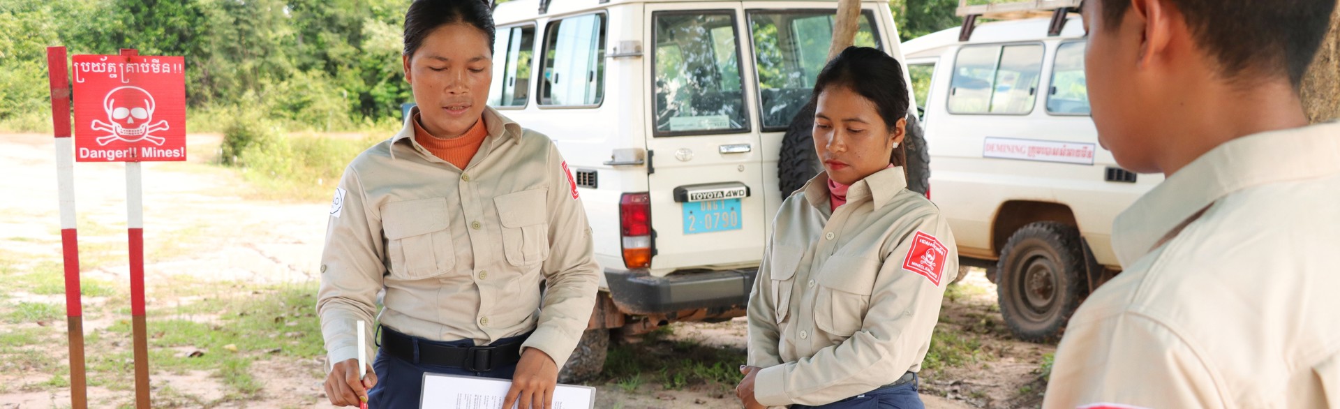 Task supervisor, Ly Sreyneang and unit commander, Pat Liv, stand behind tables with mine warning signs behind them